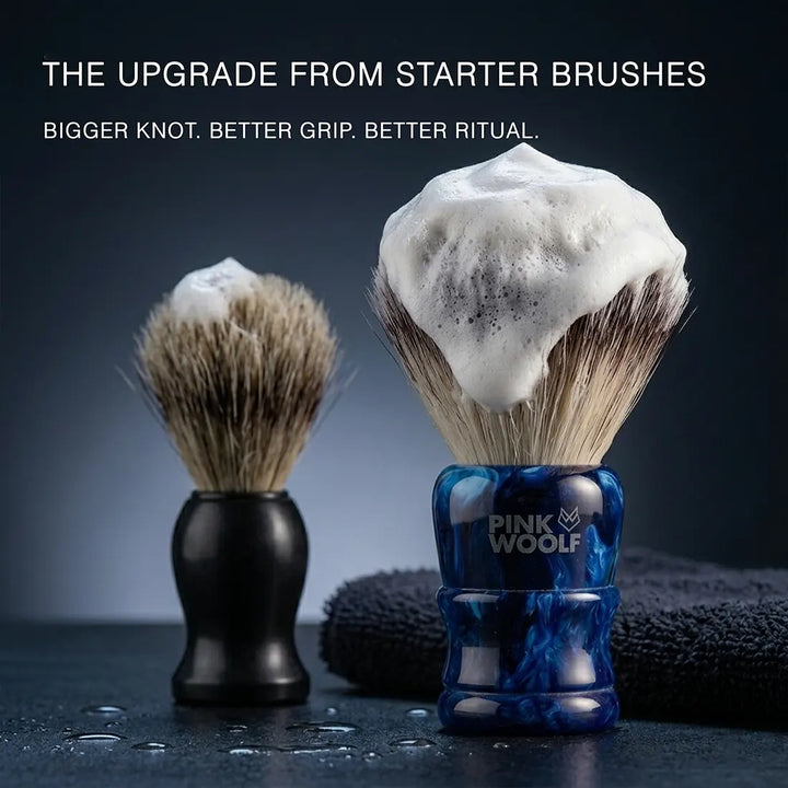 Two shaving brushes shown on a dark surface with shaving foam on the bristles. The larger foreground brush has a blue marbled handle and the text 'PINK WOOLF' is visible on the handle.
